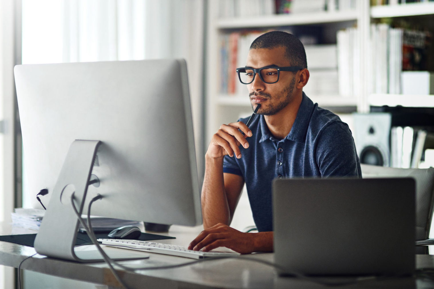 Man thinking in blue shirt with black glasses on sitting at laptop.